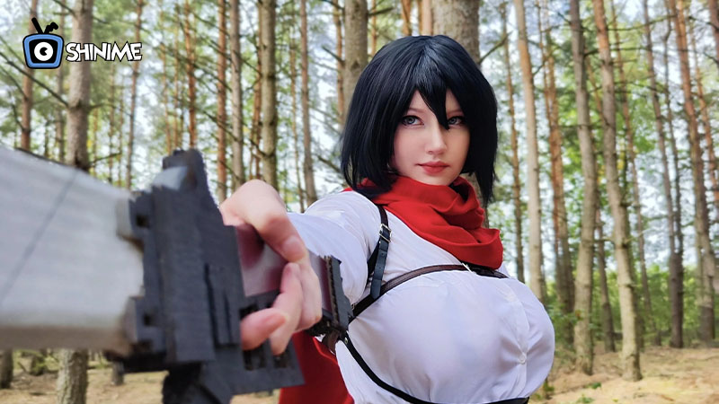 Cosplayer de Mikasa Ackerman con capa roja y equipo 3D en bosque, personaje fuerte para cosplay anime en 2025.